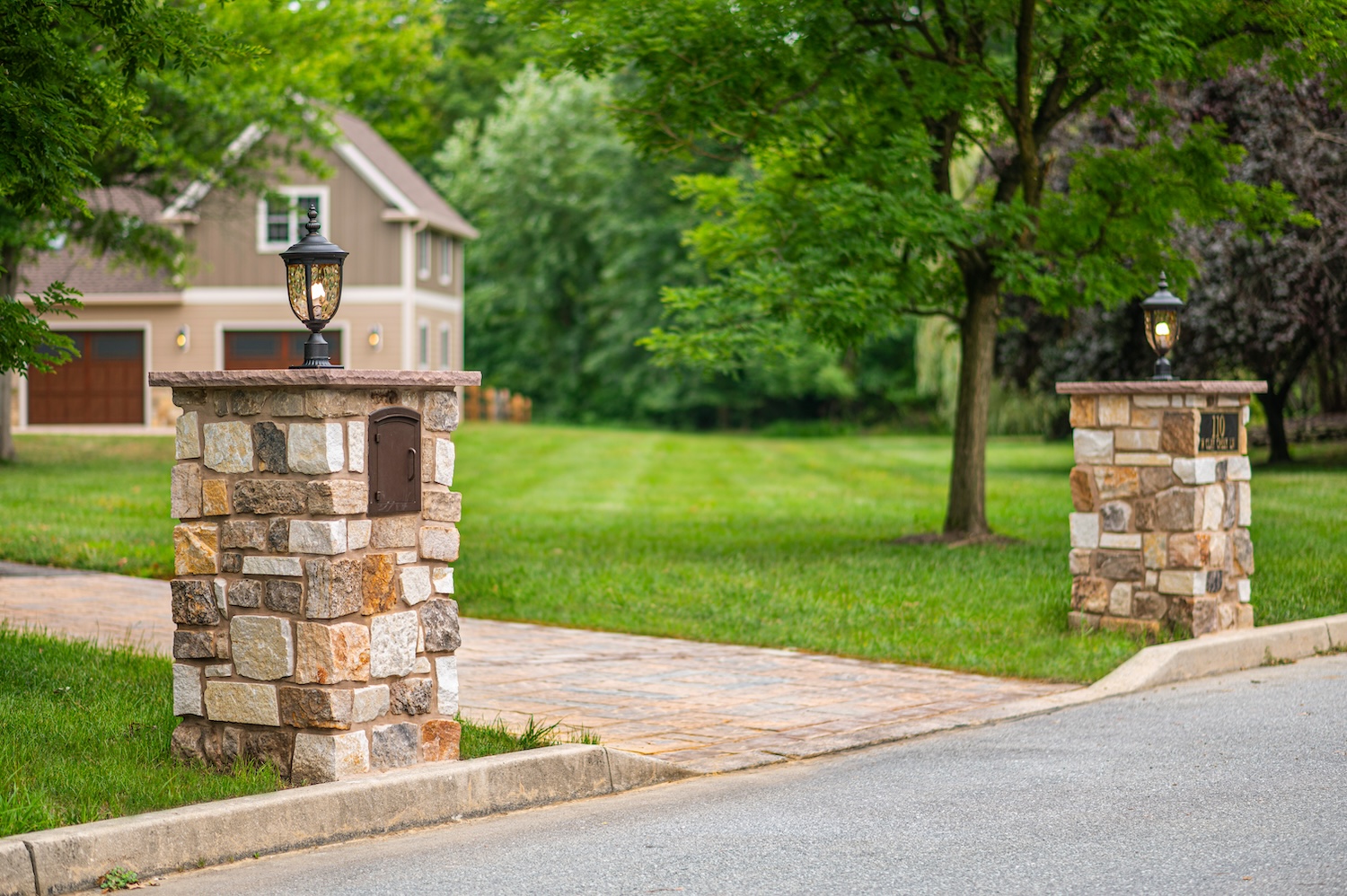 Stone pillars with lanterns stand at a driveway entrance, featuring a mailbox on one pillar. The background includes a well-maintained grassy area and a house partially visible.