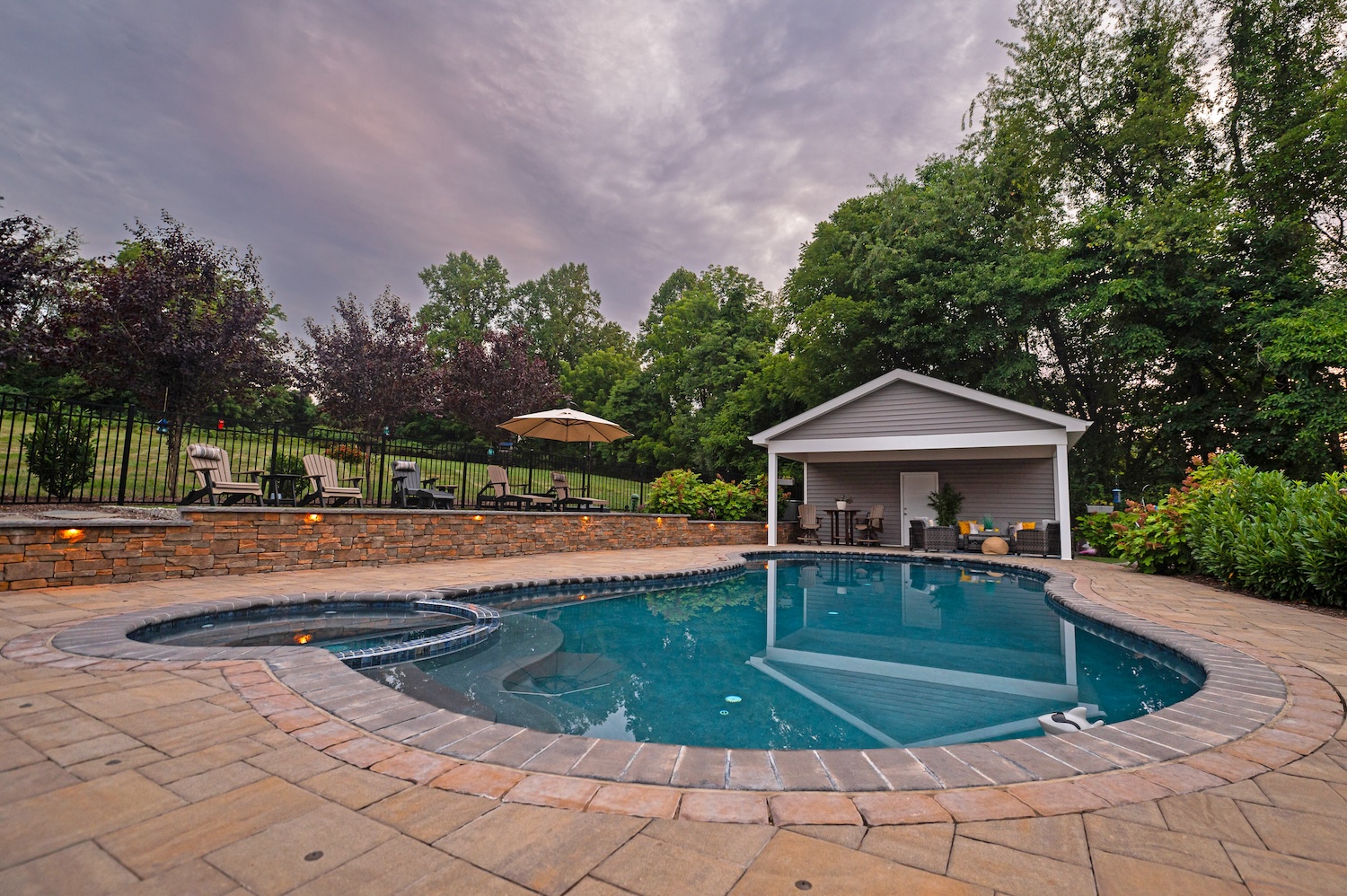 An oval swimming pool is surrounded by a stone patio. Nearby, lounge chairs face a shaded area with a table and chairs, set against a backdrop of lush greenery and a cloudy sky.