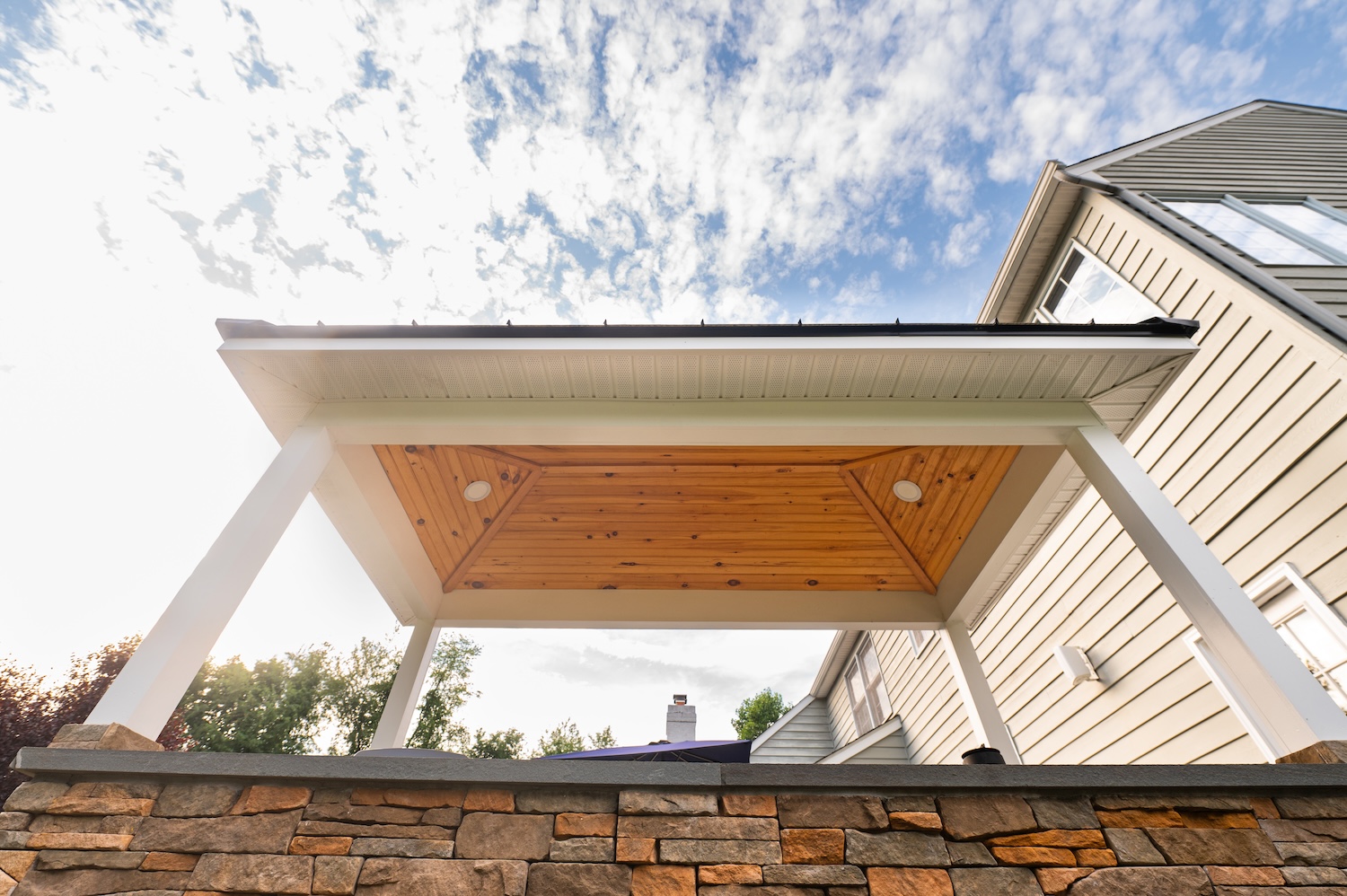 A covered patio features wooden beams and white pillars, as it stands above a stone wall. A house is visible to the right under a partly cloudy sky.
