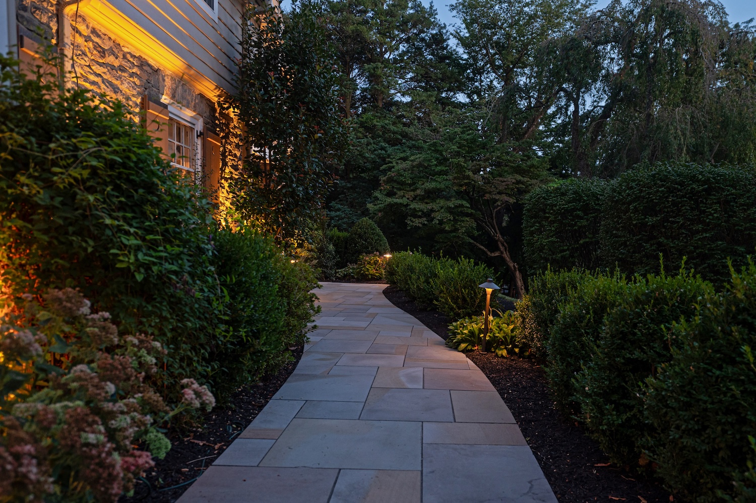A curved stone pathway, illuminated by small lights, leads through neatly trimmed hedges and shrubs alongside a stone house, set in a tranquil evening atmosphere with trees in the background.