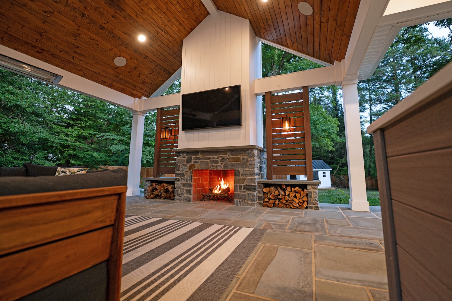 A stone fireplace with a flickering flame is the focal point, while comfortable seating and a large TV enhance the cozy outdoor space, surrounded by lush greenery and a serene atmosphere.