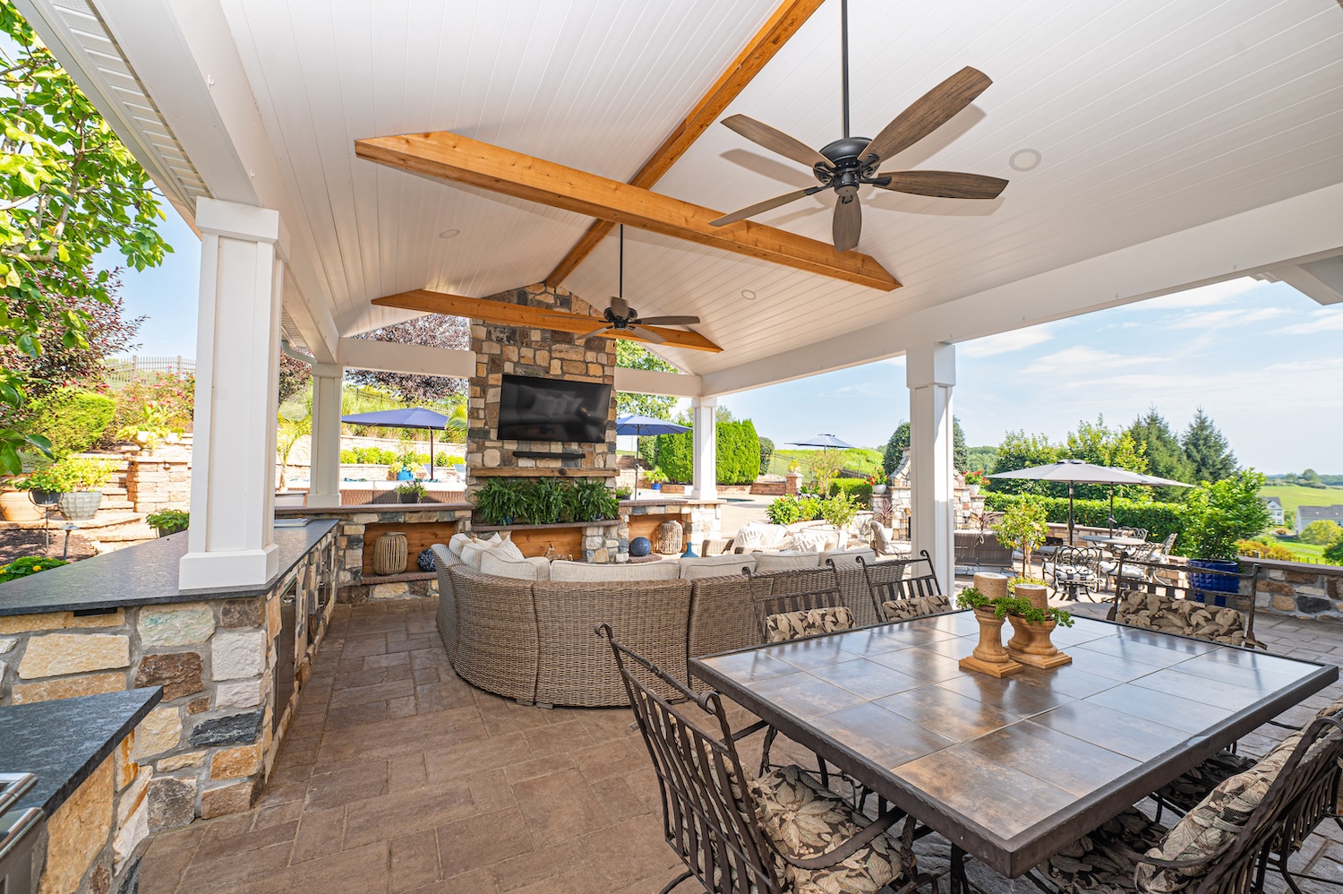 A stone-adorned outdoor patio features a circular seating area, a dining table, and ceiling fans. Lush greenery and umbrellas surround the space, creating a relaxed atmosphere for gatherings.