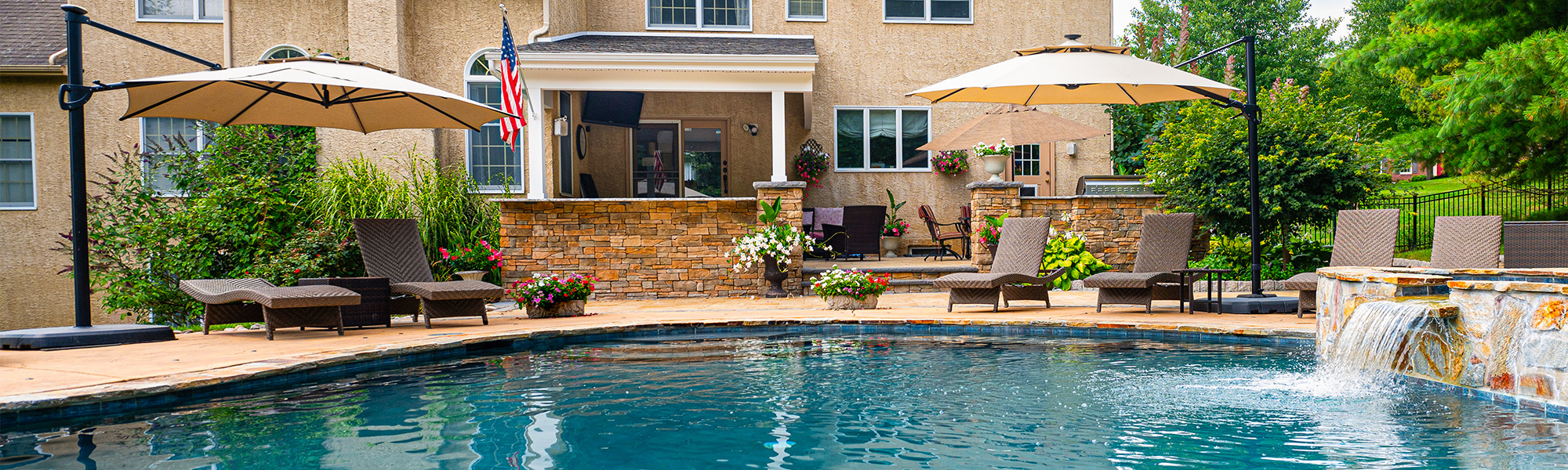 A large rectangular pool reflects the sky, bordered by stone tiles. Chaise lounges with umbrellas sit nearby, complemented by vibrant flower beds and a house with large windows in the background.