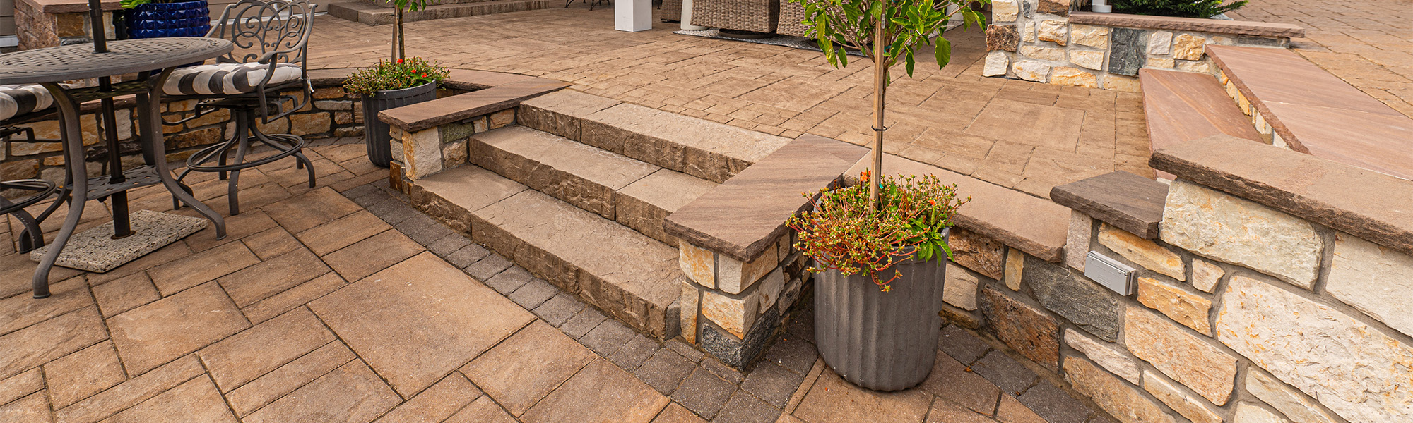 A patio features stone steps leading to a higher level, surrounded by seating with striped cushions, planters with greenery, and a textured stone wall, set in a cozy outdoor environment.