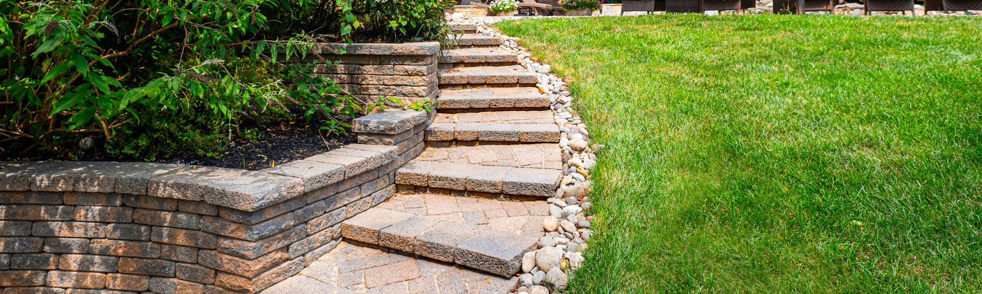 A stone pathway ascends through greenery, bordered by plants and smooth stones, leading to a grassy area with outdoor seating in the background.