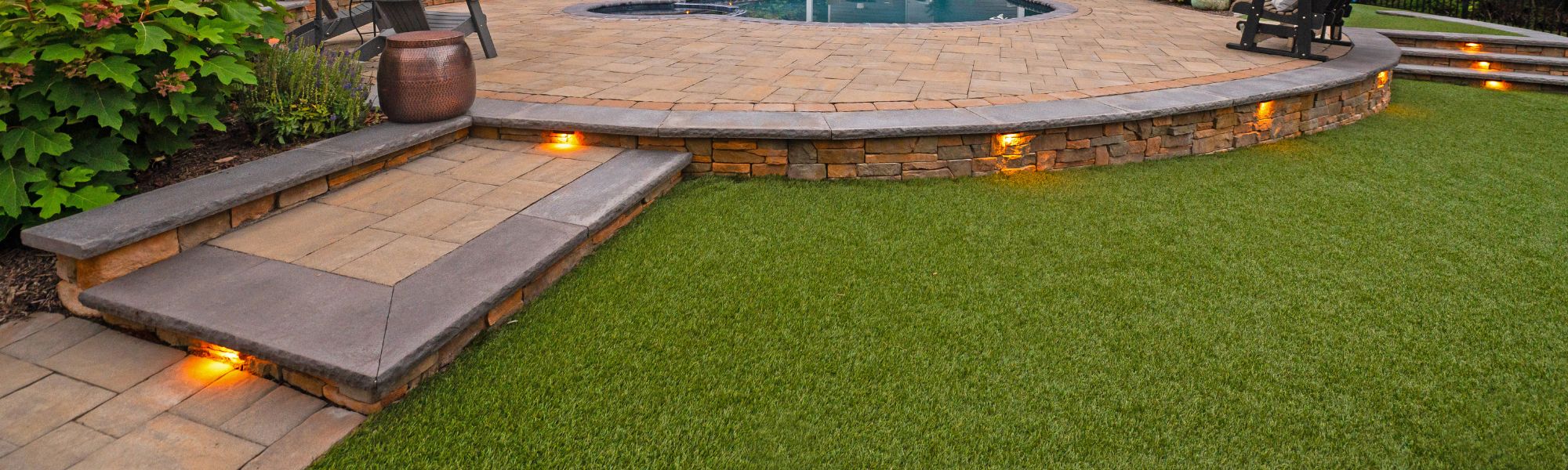 A stone patio features steps leading to a circular pool area surrounded by lush green grass, with built-in lighting illuminating the edges and vibrant plants nearby.