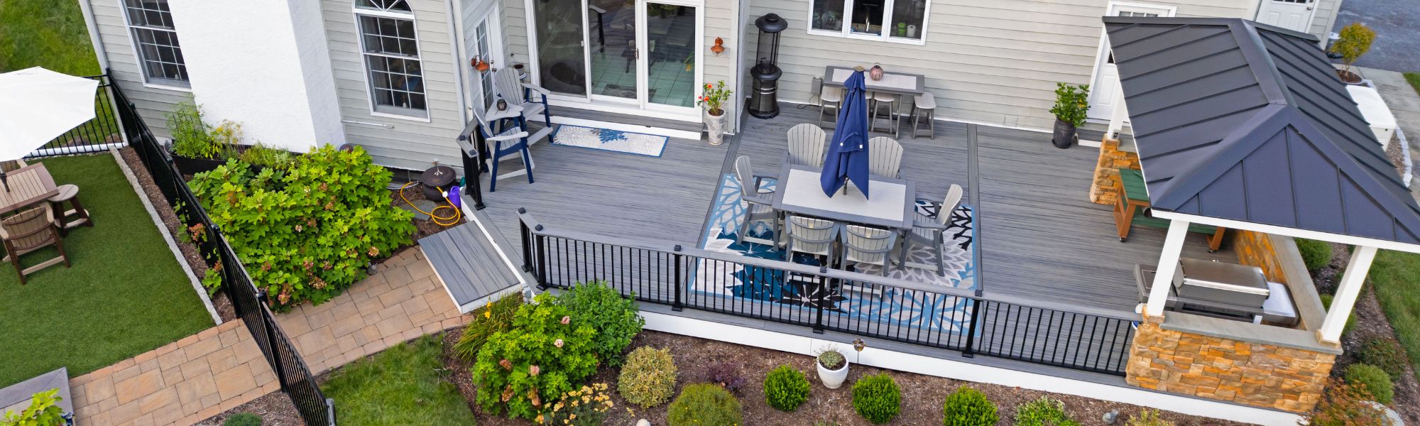 A wooden deck features a table and chairs under a large umbrella, surrounded by greenery and flower beds, with a grill and patio furniture nearby, all against a house wall.