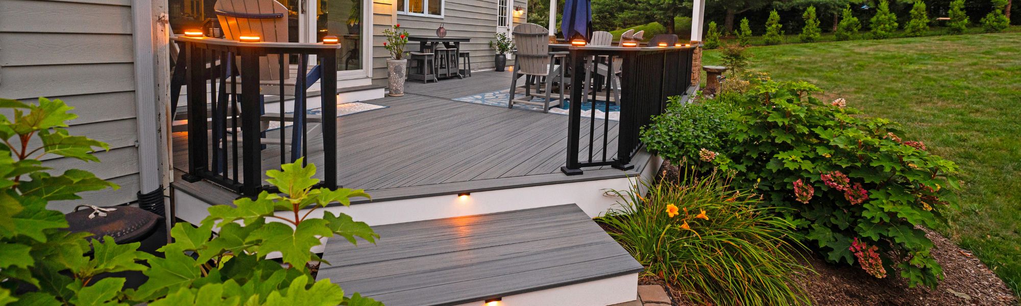 A wooden deck features steps leading up to it, with stylish outdoor furniture in a well-maintained garden. Soft lighting illuminates the space, surrounded by greenery and a grassy area.