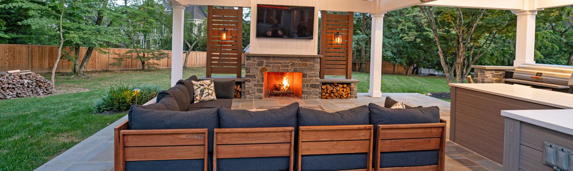 A cozy outdoor seating area features a sectional couch with dark cushions, a stone fireplace glowing with fire, and a TV mounted above it, surrounded by trees and a wooden fence.
