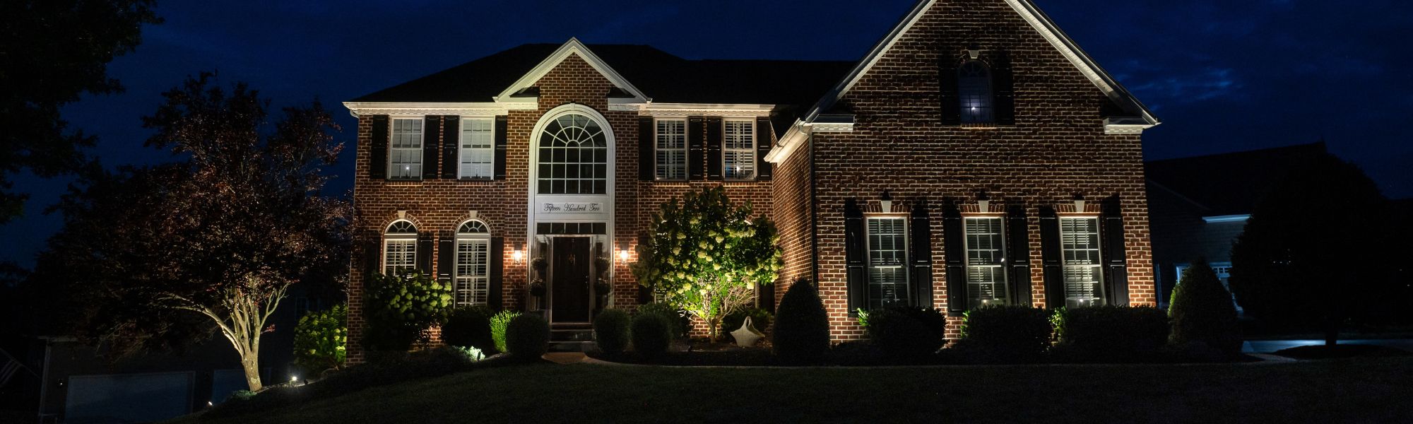 A brick house with white trim stands illuminated at night, surrounded by neatly trimmed shrubs and trees, creating a welcoming atmosphere in a quiet residential area.