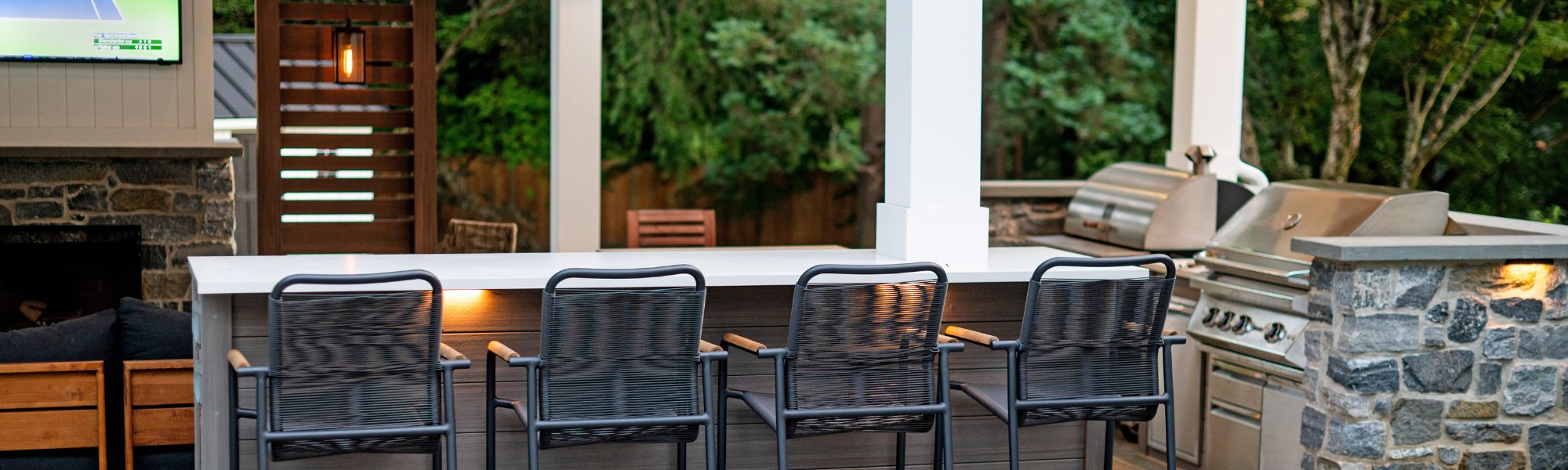 A stone patio features three black wicker chairs, facing a sleek outdoor bar. In the background, a large grill and a TV display create a relaxed, inviting atmosphere surrounded by greenery.