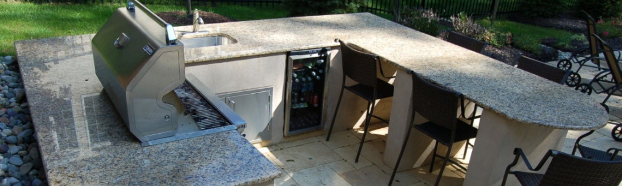 A polished granite outdoor kitchen features a built-in grill, a sink, and a beverage cooler, surrounded by stone flooring and landscaped greenery, creating a functional space for entertaining.