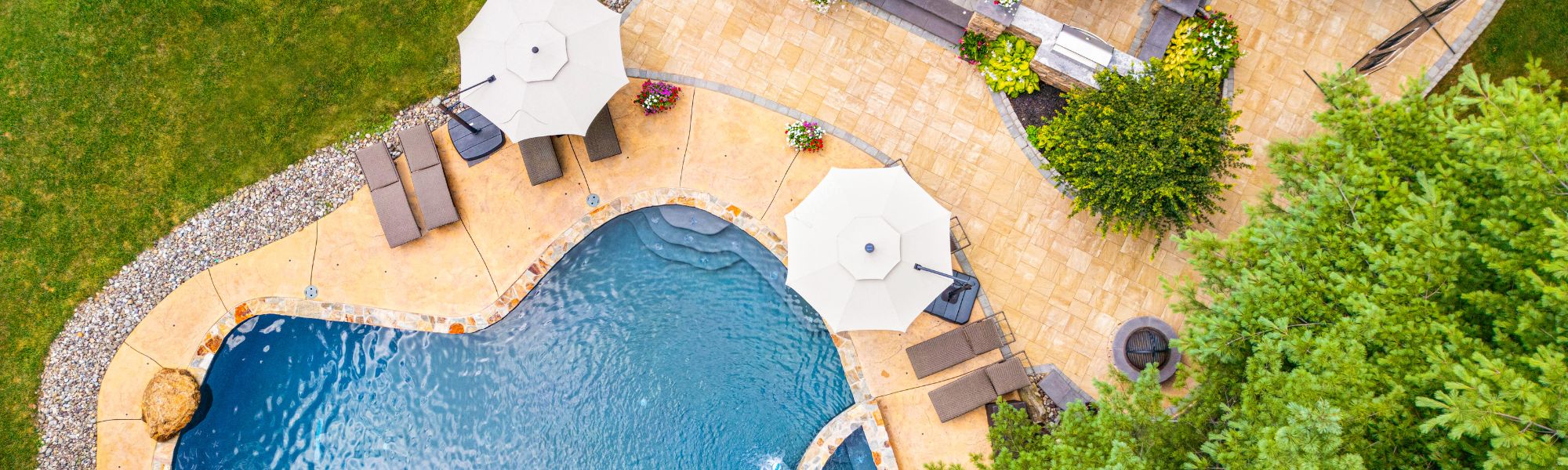 A blue pool is surrounded by tan paving stones, sun loungers, and umbrellas. Lush greenery and flowers enhance the serene outdoor setting, suggesting a relaxing leisure area.