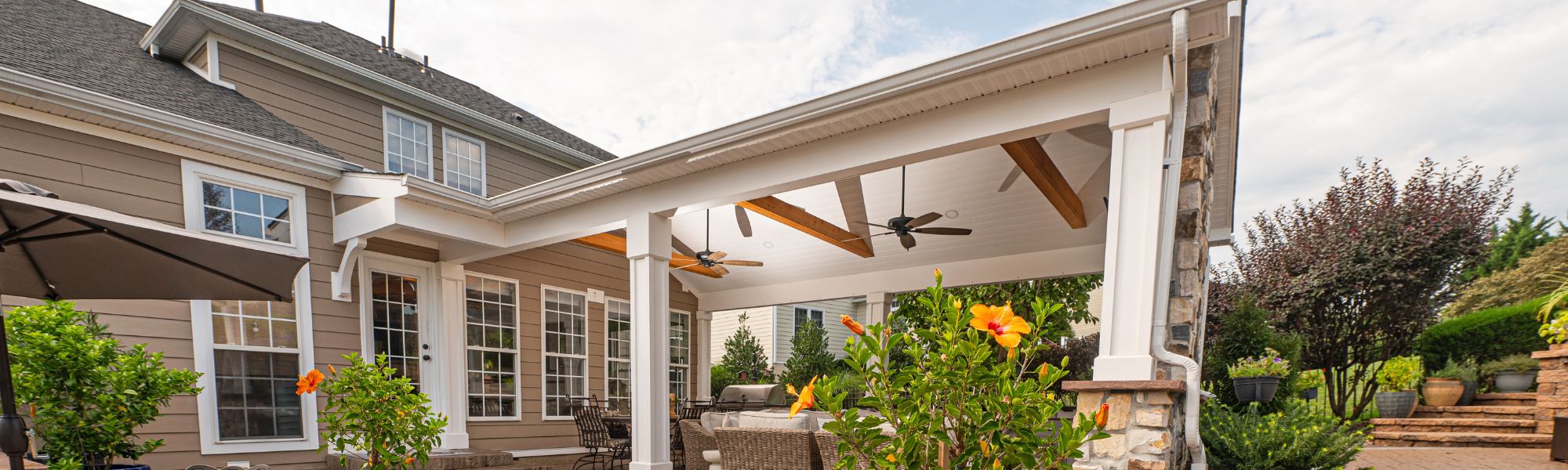 A covered patio with wooden beams features comfortable seating and tropical plants. The environment includes a landscaped yard with shrubs and flowers, under a partly cloudy sky.