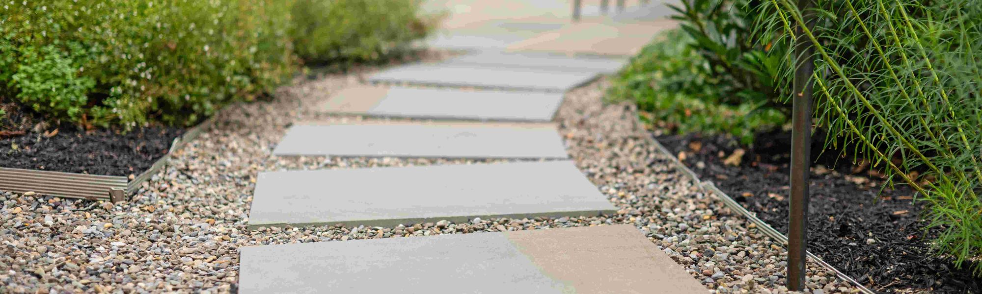A pathway made of concrete stepping stones is placed on a bed of small pebbles, surrounded by green shrubs and a black mulch area, leading through a garden space.