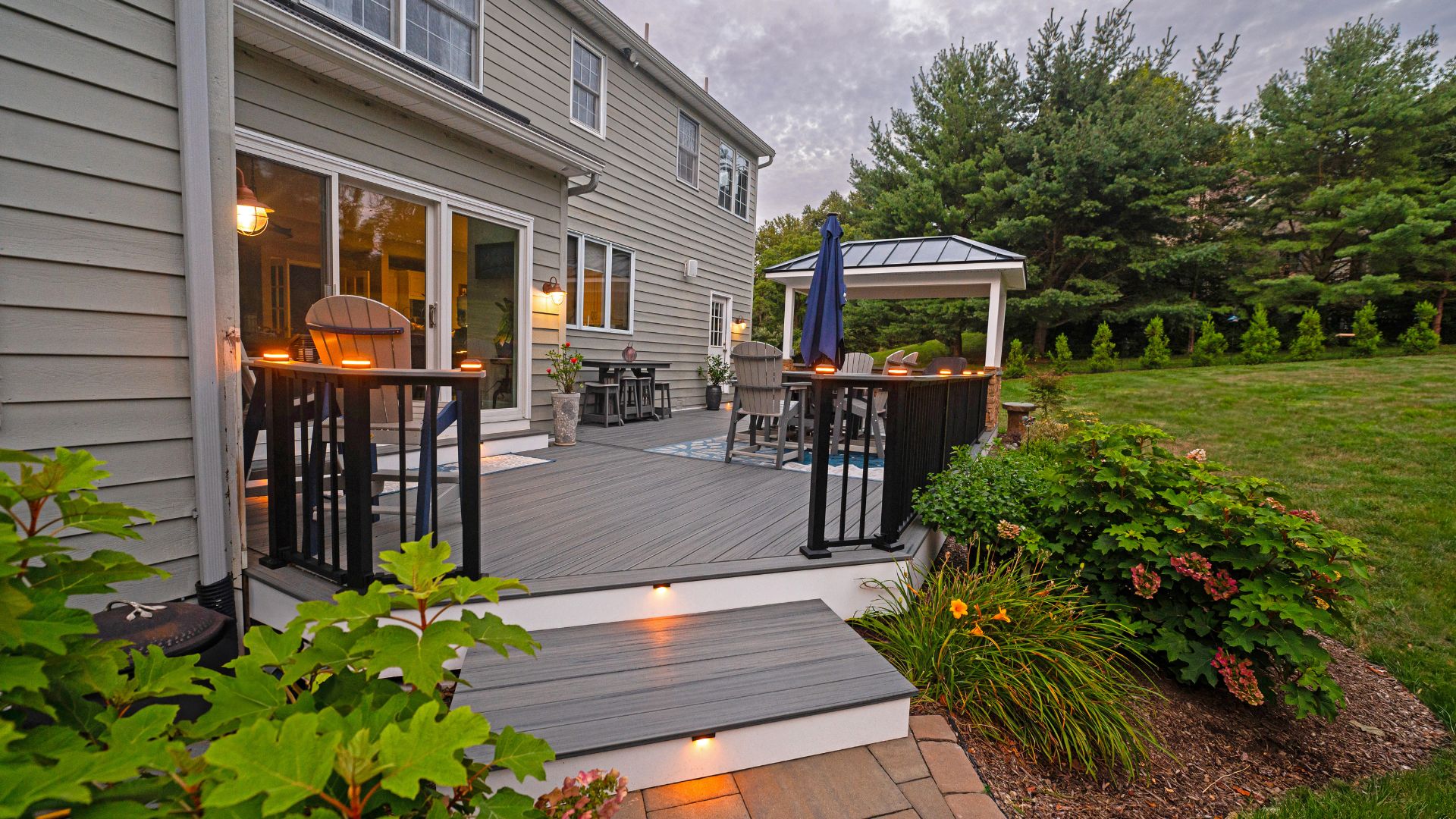 A wooden deck featuring outdoor chairs and a table is illuminated by soft lights. It is framed by lush greenery and a gazebo-like structure, set against a cloudy sky.