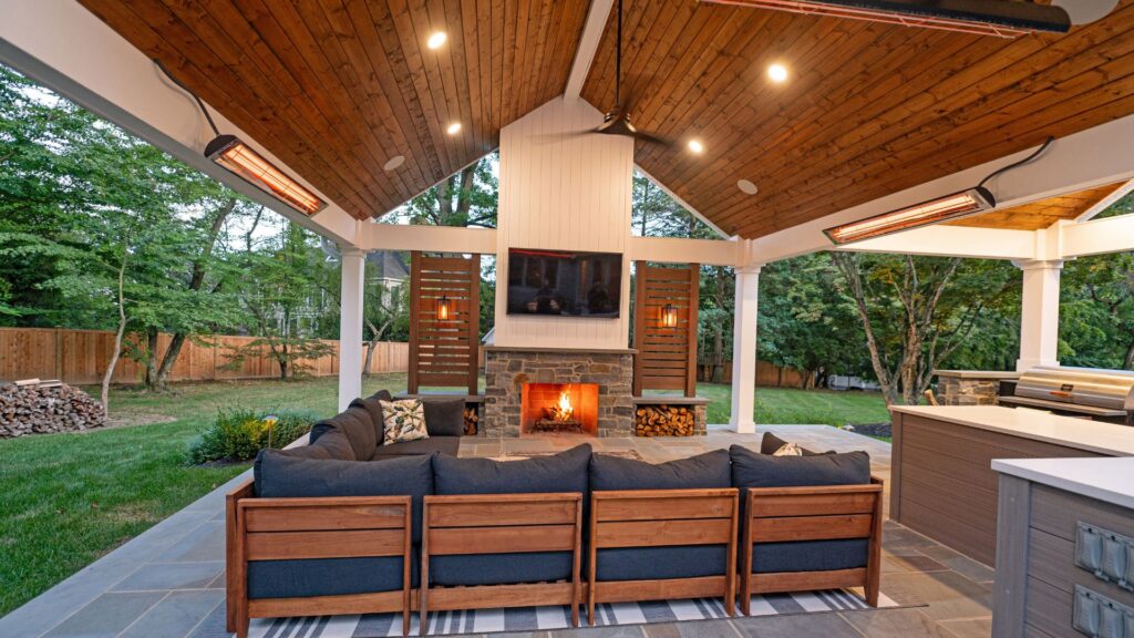 A cozy outdoor living area features a sectional sofa facing a stone fireplace, illuminated by ceiling lights. A television hangs above the fireplace, surrounded by lush greenery and a wooden fence in the background.