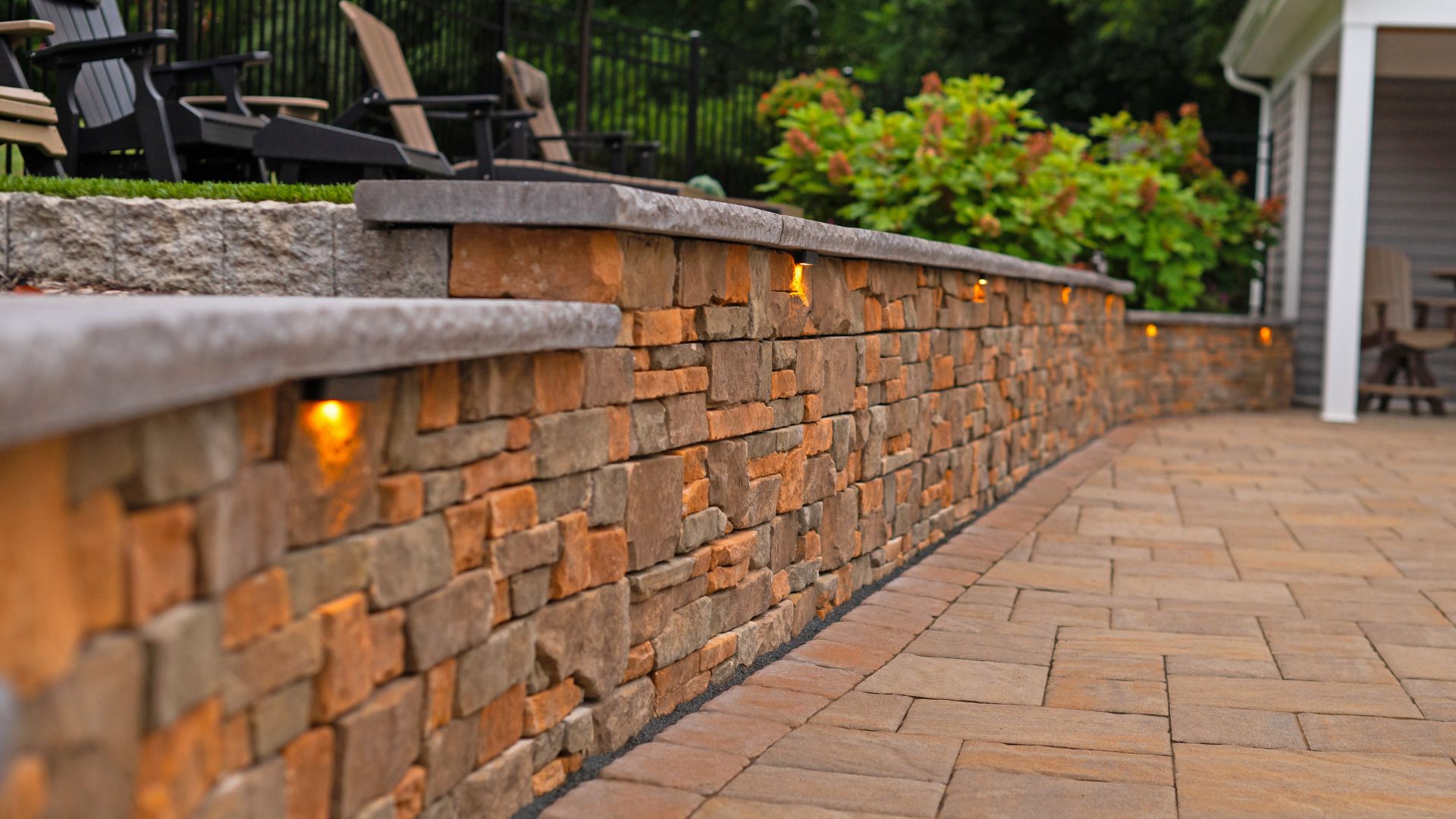 A stone wall with integrated lighting sits along a paved pathway. Chairs are visible in the background, surrounded by greenery in a residential outdoor space.