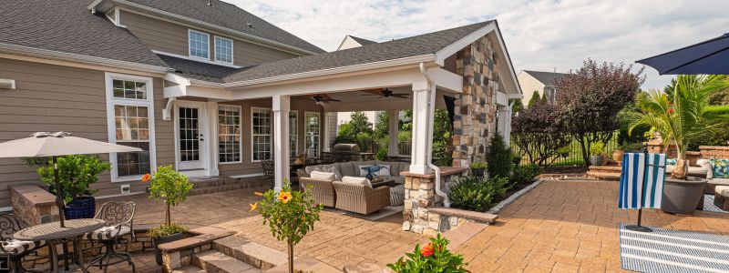 A stylish patio features comfortable seating under a roof, surrounded by landscaped greenery. Sunlight filters through the area, creating an inviting outdoor space for relaxation and socializing.