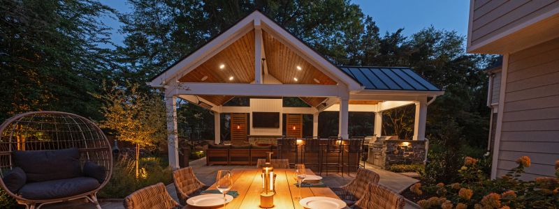A wooden dining table is set with plates and candles, surrounded by a cozy outdoor gazebo. Soft lighting illuminates the space, creating a warm atmosphere in a lush garden setting.