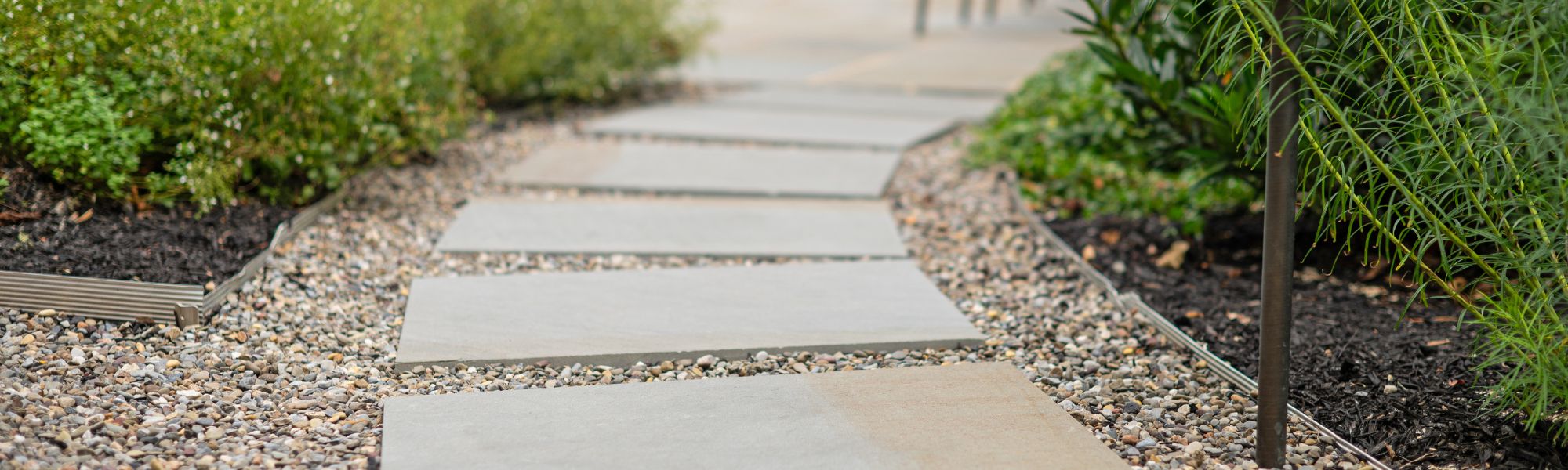 Square stone slabs create a walking path, positioned amidst gravel and bordered by green foliage, leading into a blurred background of more landscaping elements.
